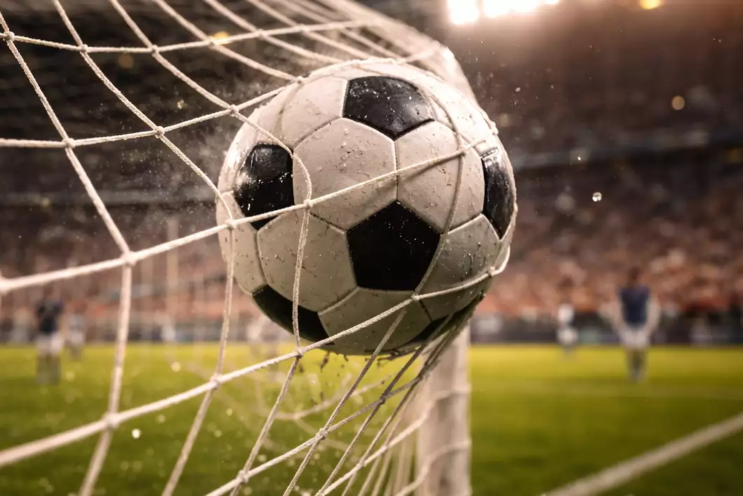 Balón de fútbol entrando en la portería sobre césped natural durante un partido nocturno iluminado por focos